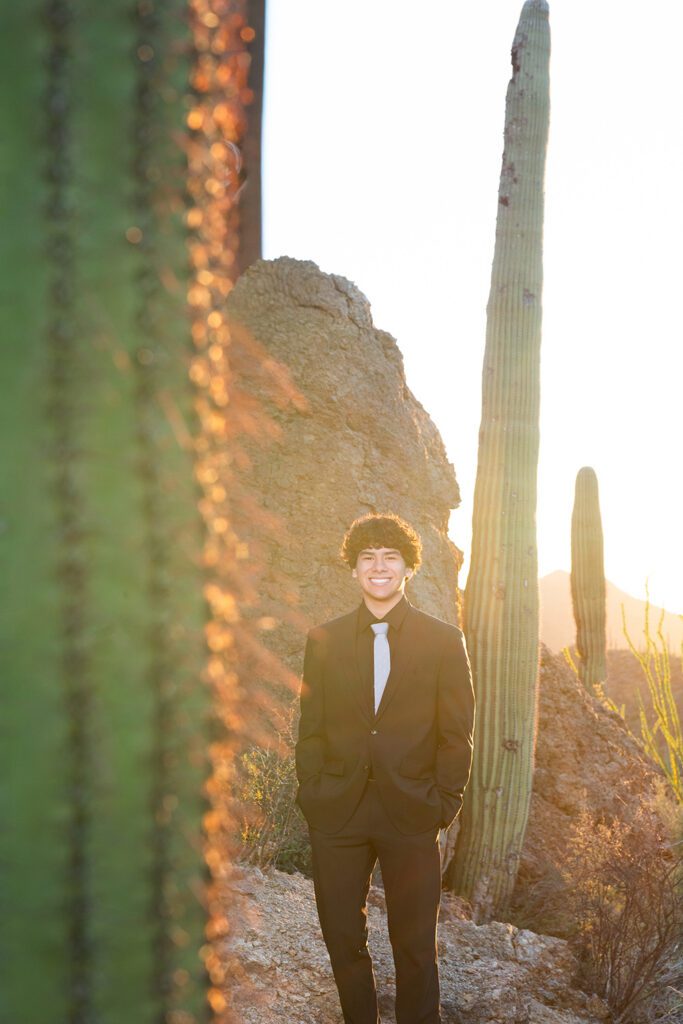High school senior guy standing on rocky overlook at Gates Pass in Tucson wearing black suit at sunrise with saguaro cactus and mountain views