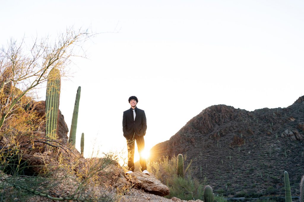 High school senior guy standing on rocky overlook at Gates Pass in Tucson wearing black suit at sunrise with saguaro cactus and mountain views
