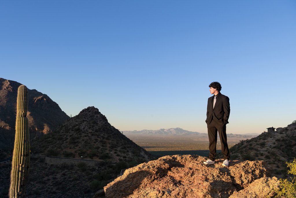 High school senior guy standing on rocky overlook at Gates Pass in Tucson wearing black suit at sunrise with saguaro cactus and mountain views